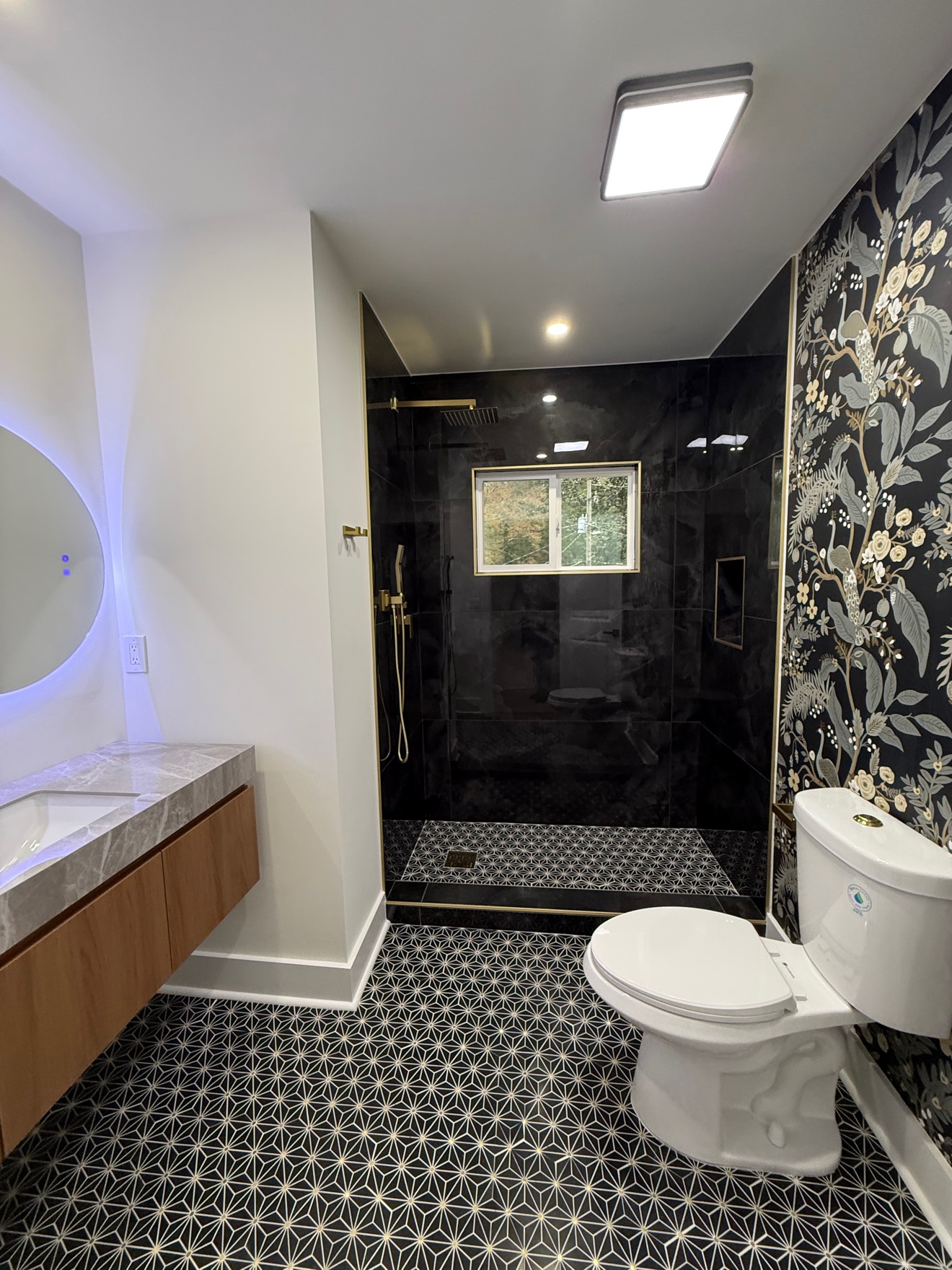 Bathroom with a black tile walk-in shower, gold fixtures, floral wallpaper and geometric floor tile