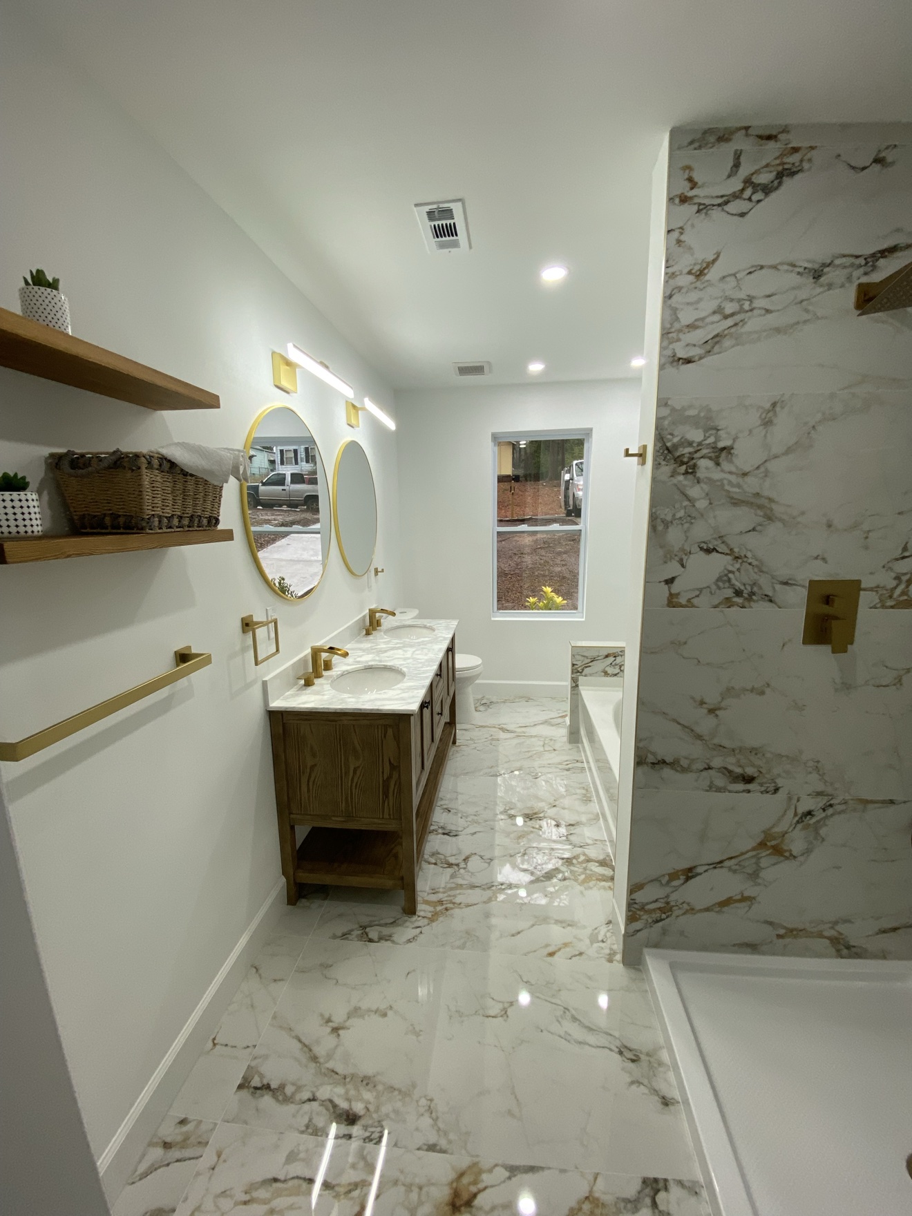 Marble-tile bathroom with a double wood vanity, two round gold mirrors and gold faucets